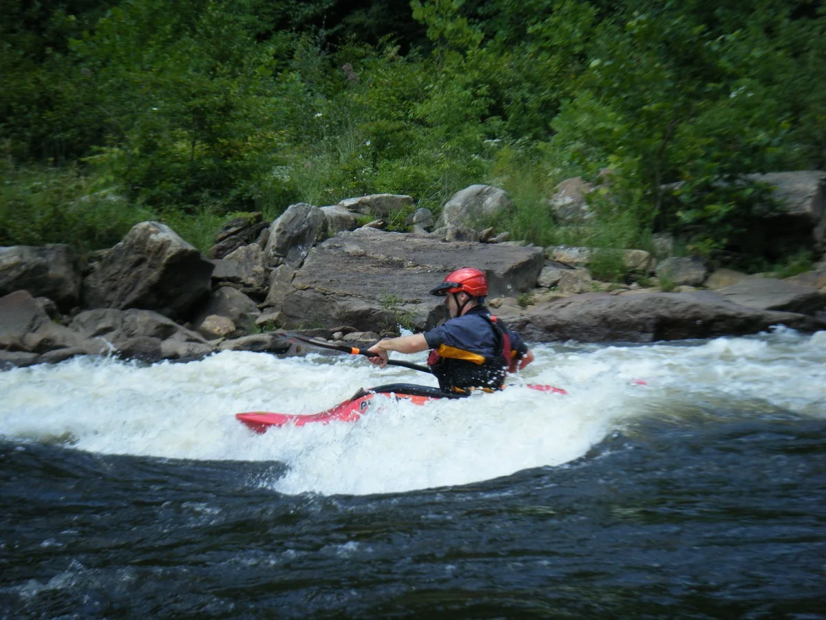 A kayaker paddles through whitewater beside a wooded riverbank.
