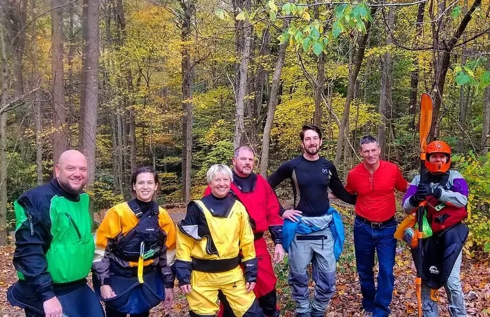 A group of paddlers stands together beside a wooded riverbank.