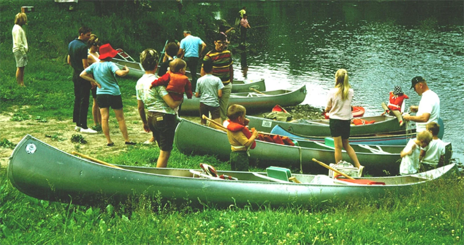 People gather with canoes on a grassy riverbank.