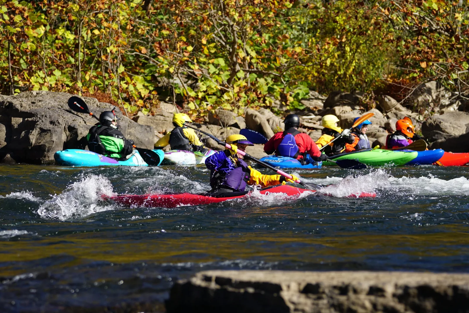 Kayakers paddle through a rocky river in autumn.